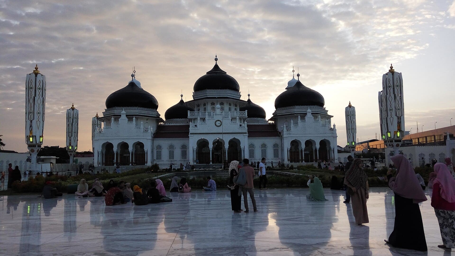 The Baiturrahman Grand Mosque in Banda Aceh at dusk, its white arcades and black domes above a wet marble forecourt where people sit and walk