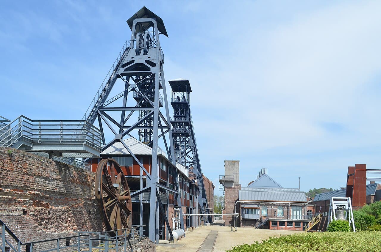 The two steel headframes of the Bois du Cazier colliery at Marcinelle rising over the pithead, with a rusted winding wheel and brick buildings below them under a blue sky