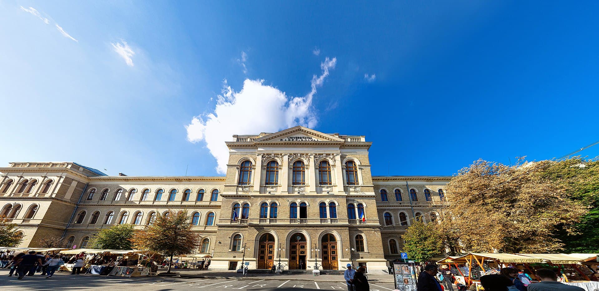 The main building of Babeș-Bolyai University in Cluj-Napoca, its pedimented centre and long brick wings seen from the street, with market stalls trading in front