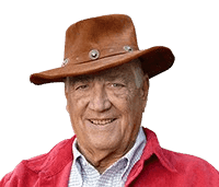 An elderly man wearing a brown leather cowboy hat and a red jacket smiles warmly at the camera. He stands in front of a wooden fence with greenery in the background, giving the image an outdoorsy, rustic feel. His friendly expression and casual attire convey a relaxed, approachable vibe.