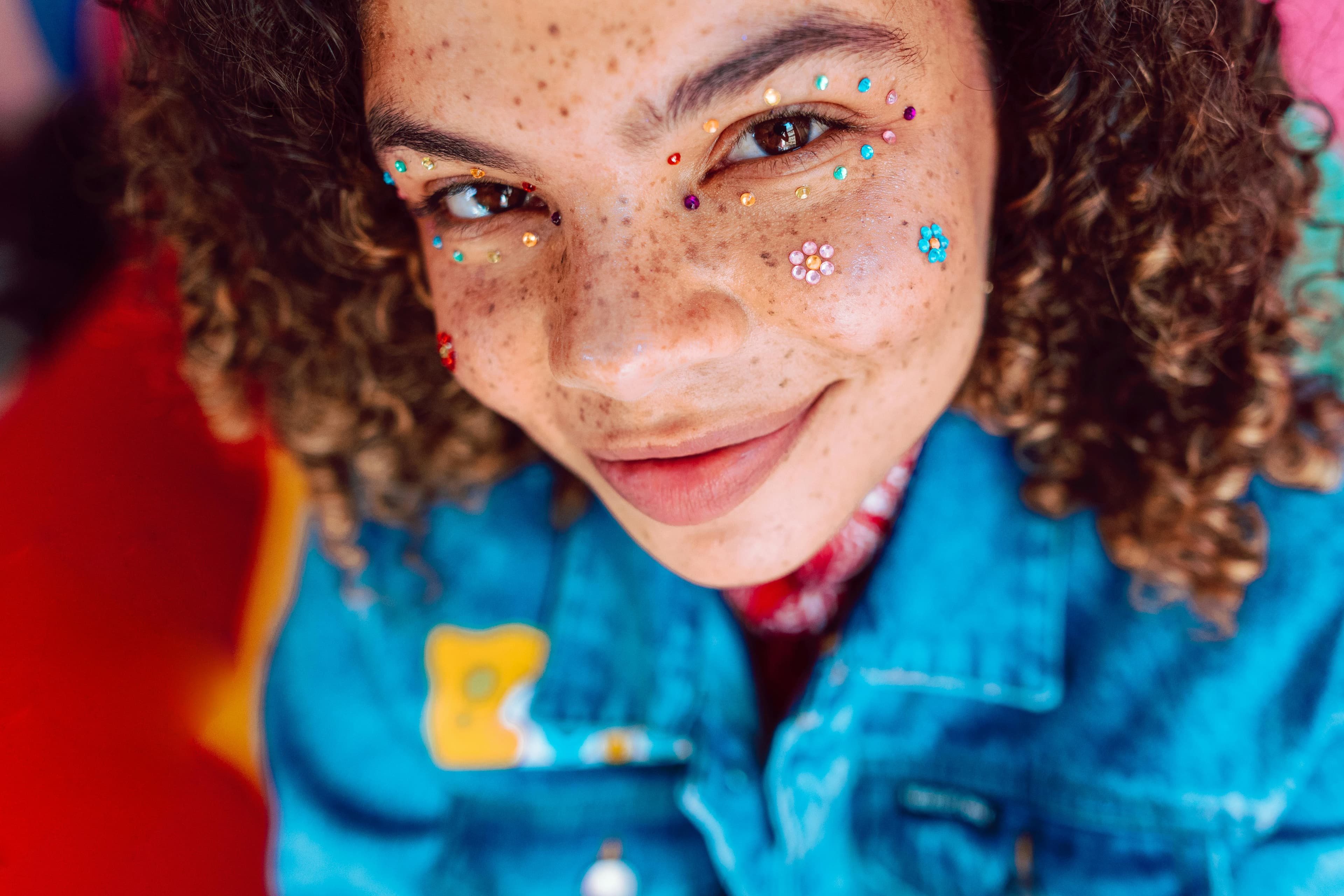 A cheerful young woman with freckles and curly hair adorned with colorful rhinestones and floral face decorations, wearing a vibrant denim jacket. This close-up portrait radiates creativity, self-expression, and festival-inspired style.
