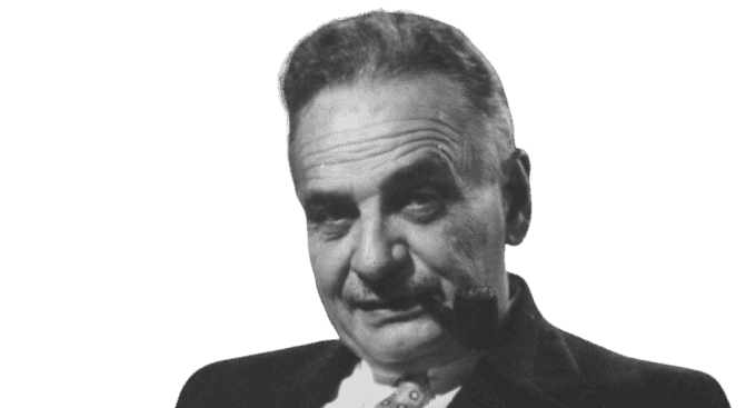 A black-and-white portrait of an older man with a pipe in his mouth, giving a contemplative expression. He has short, combed-back hair with a receding hairline, and is dressed in a suit jacket and collared shirt. The background includes shelves of books, suggesting an intellectual or scholarly setting.