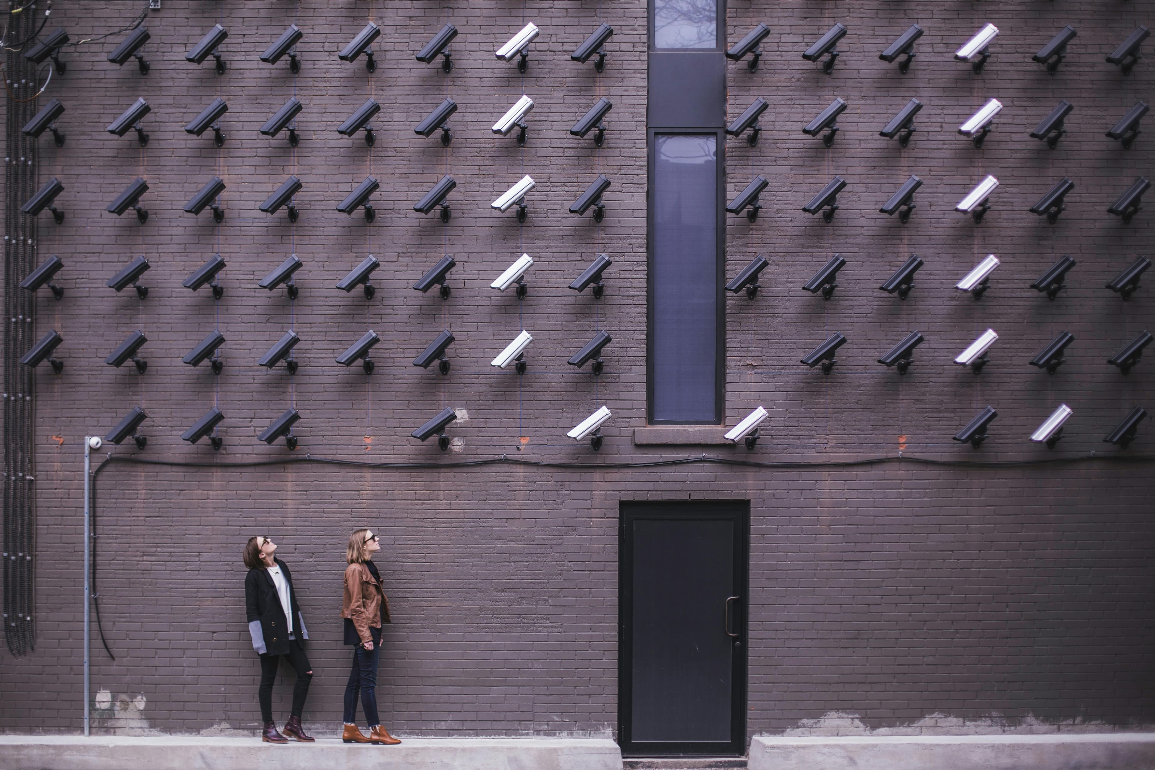 Two people stand in front of a brick wall covered with numerous surveillance cameras, all pointed in various directions. This visually striking scene evokes themes of surveillance, privacy, and modern societal control in an urban environment.