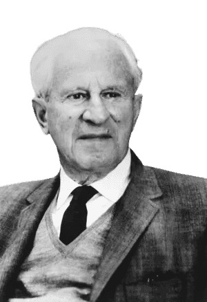 Black and white portrait of an elderly man with short, white hair, dressed in a suit jacket over a sweater and tie. He is seated in front of a bookshelf, gazing slightly to the side with a thoughtful expression. His formal attire and the background suggest an intellectual environment, conveying a sense of wisdom and scholarly presence.