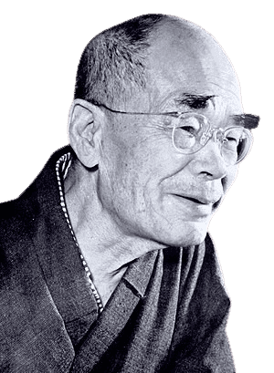 Black and white portrait of an elderly man with a shaved head, round glasses, and a gentle smile. He is dressed in traditional clothing with a visible patterned collar. His expression is serene and thoughtful, suggesting wisdom and a reflective demeanor.