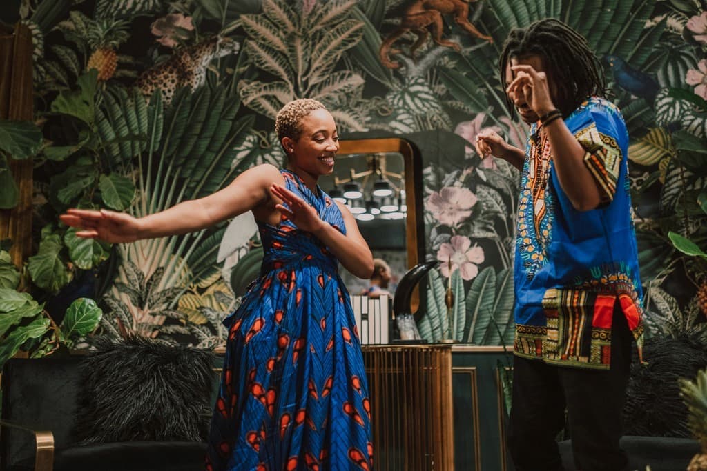 A joyful couple dancing in a vibrant, plant-themed room, both wearing colorful African-inspired attire, showcasing their cultural pride and the lively ambiance of their surroundings.