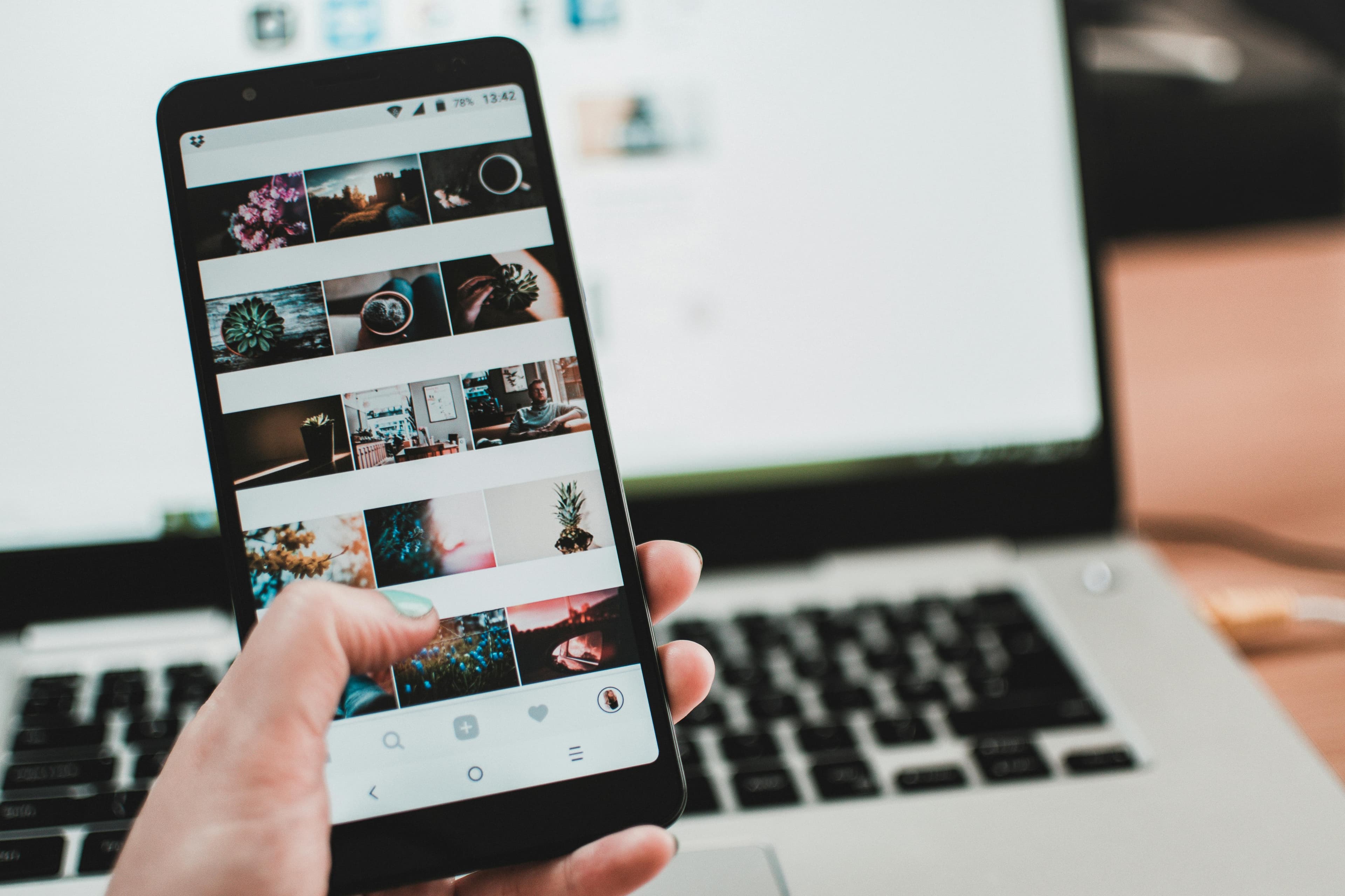 A hand holding a smartphone displaying a photo gallery app, with a blurred laptop screen in the background. This image illustrates multitasking, digital content browsing, and the integration of mobile and desktop technology in everyday life.
