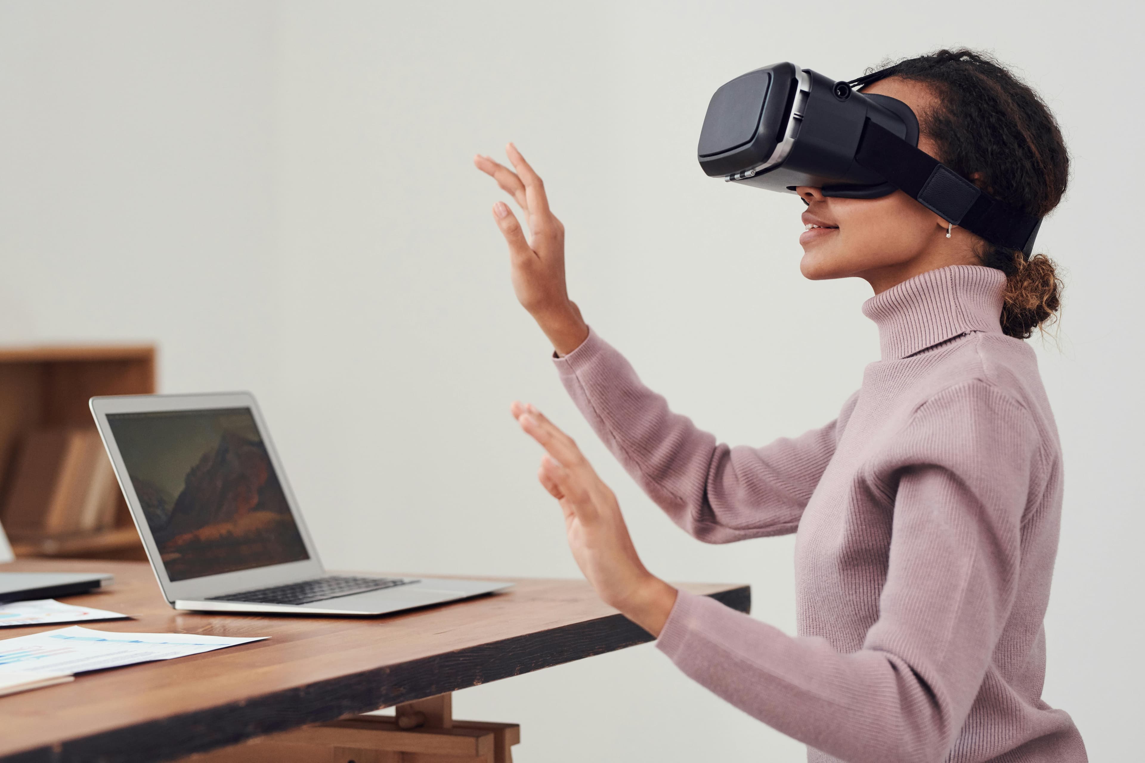 A woman wearing a virtual reality (VR) headset interacts with a virtual environment, gesturing with her hands, while sitting at a desk with an open laptop in the background. This image highlights modern technology, VR applications, and remote or digital work setups.