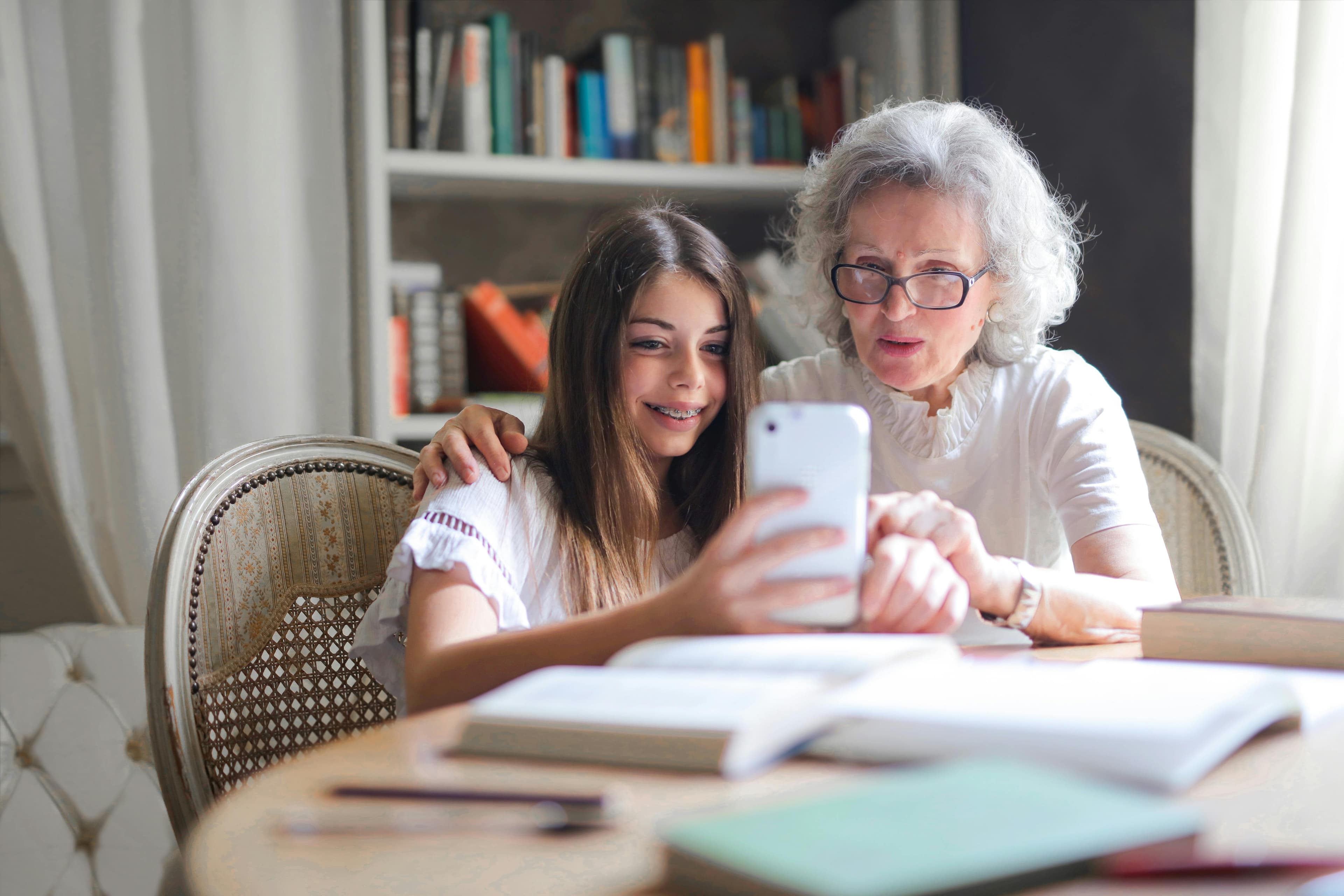 A grandmother and her granddaughter sit together at a table, smiling while looking at a smartphone, surrounded by open books. The scene highlights the blending of generations, technology, and shared learning experiences in a warm, inviting atmosphere.