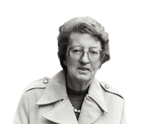A black-and-white photograph of an elderly woman with glasses and short, wavy hair, wearing a trench coat and a pearl necklace. She stands on a city street, giving a composed expression, evoking a sense of sophistication and resilience. The urban background suggests a mid-20th-century setting.