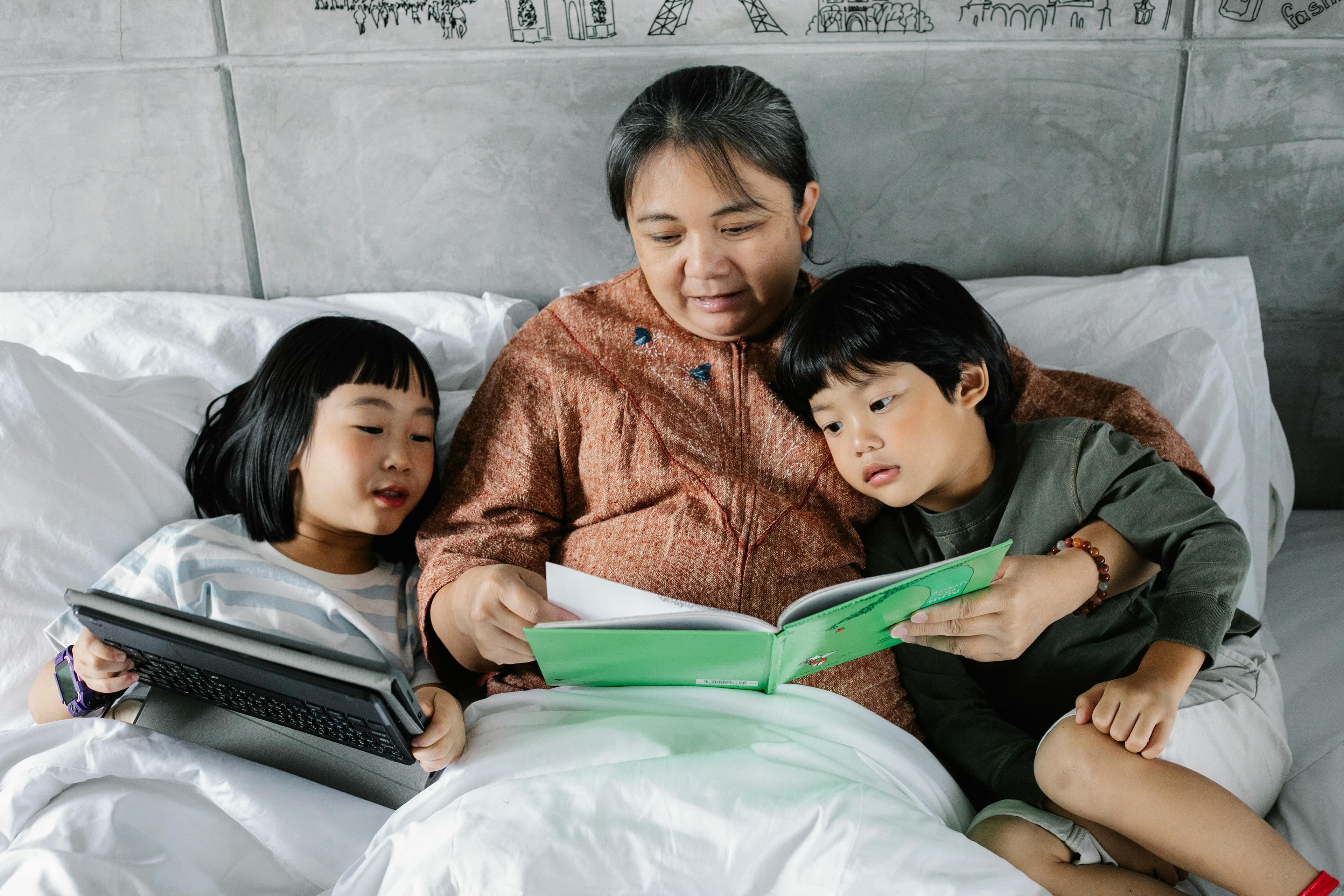 A grandmother sits in bed with two young children, reading a book together while one child holds a tablet. The cozy scene highlights family bonding, storytelling, and the blend of traditional and modern learning methods.
