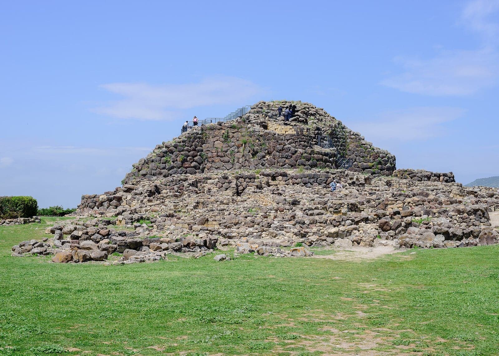 The Bronze Age nuraghe of Su Nuraxi at Barumini in Sardinia, a broad tower of dark dry-stone blocks rising above the excavated stone huts of the village around it
